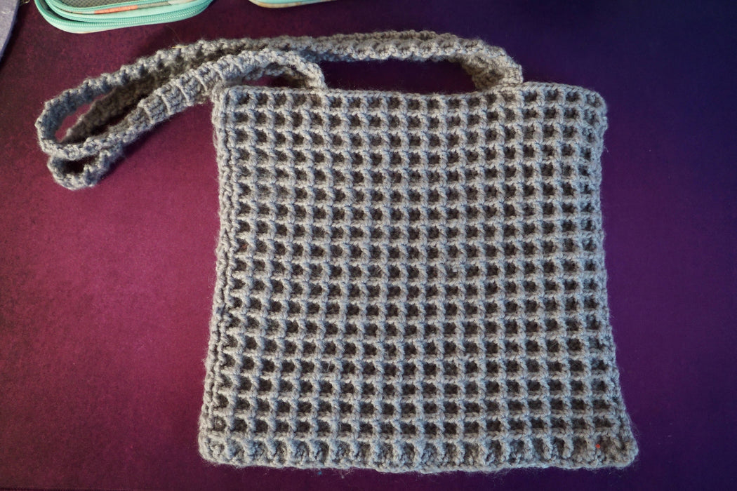 A grey square bag crocheted with a waffle-shaped stitch pattern, made with two sides attached together. Each side has its own strap made from the same yarn.